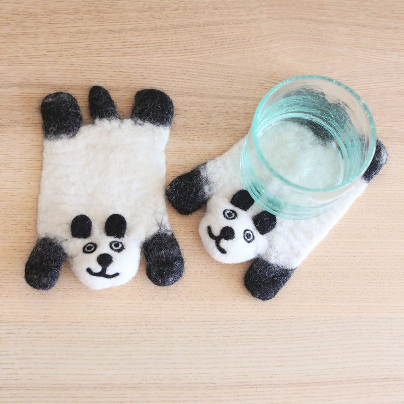 Two handcrafted felt panda coasters rest on a light wooden surface; one flat and one beneath a clear glass. Each charming wool felt panda coaster is primarily white with black accents on its ears, paws, and facial features, including a small smiling mouth and simple eyes. Crafted from soft 100% sheep wool, their handmade details create a warm, inviting charm, highlighting their ethical production by Nepalese female artisans.