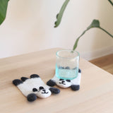 Two handmade Felt Panda Coasters are displayed on a light wooden table, one holding a clear, light blue-tinted glass of water, with a blurred green plant in the background. Each charming panda coaster is crafted from soft white sheep wool felt, with black felt accents forming their ears, paws, and playful facial features. These ethically handmade wool felt coasters offer both whimsical decoration and practical use, supporting female artisans.