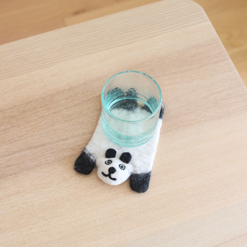 A light blue glass rests on a charming white felt panda coaster, part of a set, displayed on a light wood surface. This handcrafted wool felt panda coaster features a soft white body with black ears, legs, and a cute facial design. These ethically handmade felt panda coasters bring a playful touch to any home decor.