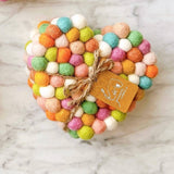 A single pastel circle felt coaster, part of a set, rests on a marbled surface. This handcrafted wool felt coaster is ethically made from small, hand-stitched organic lamb wool felt balls, supporting female artisans. The approximately 4-inch diameter coaster features soft pastel hues of pink, green, orange, blue, and white, with a brown tag tied by jute string.