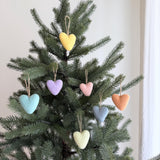 Seven pastel felt heart ornaments are showcased on a lush green Christmas tree, evoking a feeling of warmth and handmade charm. These unique Christmas ornaments, crafted from needle-felted wool felt, display soft pastel shades including yellow, light purple, pink, light blue, orange, grey, and light green. Each heart-shaped wool decoration features a rustic jute string for hanging.