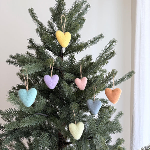 Seven pastel felt heart ornaments are showcased on a lush green Christmas tree, evoking a feeling of warmth and handmade charm. These unique Christmas ornaments, crafted from needle-felted wool felt, display soft pastel shades including yellow, light purple, pink, light blue, orange, grey, and light green. Each heart-shaped wool decoration features a rustic jute string for hanging.
