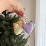 Seven handmade wool felt pastel heart ornaments are delicately held by a female hand against a lush green Christmas tree. Each needle-felted heart-shaped ornament, crafted from soft sheep's wool, displays gentle pastel hues of peach, yellow, lavender, grey, and green. A rustic jute string is attached to each unique Christmas ornament for hanging.