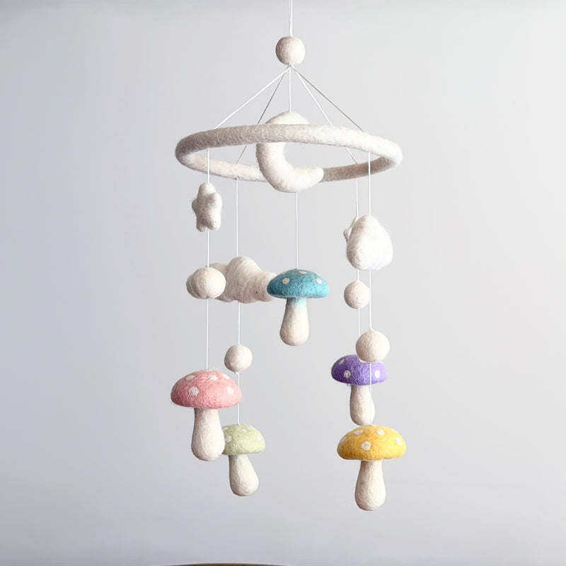 A Felt Pastel Mushroom Baby Mobile hangs against a pale gray backdrop, creating a whimsical and dreamy scene. This charming wool felt baby mobile features pastel mushrooms in pink, light green, blue, purple, and yellow, each with white polka dots. Fluffy white wool felt stars, clouds, and a gentle crescent moon dangle at varying lengths from a soft white felt ring, suspended by thin white strings.