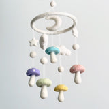 A Felt Pastel Mushroom Baby Mobile is centered against a pale background, creating a calm and whimsical atmosphere. It features a circular wool felt frame from which various soft wool felt shapes are suspended. These include five delicate pastel mushrooms in shades of purple, blue, green, pink, and yellow with white dots, complemented by fluffy white wool felt clouds, stars, and a gentle crescent moon.