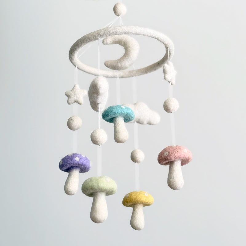A Felt Pastel Mushroom Baby Mobile is centered against a pale background, creating a calm and whimsical atmosphere. It features a circular wool felt frame from which various soft wool felt shapes are suspended. These include five delicate pastel mushrooms in shades of purple, blue, green, pink, and yellow with white dots, complemented by fluffy white wool felt clouds, stars, and a gentle crescent moon.