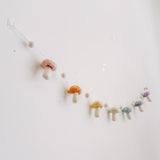 A felt pastel mushroom garland is displayed on a light, neutral wall, evoking a whimsical and cozy atmosphere. The handcrafted felt garland is attached with transparent hooks and features alternating soft pastel wool felt balls and wool felt mushrooms. Each wool felt mushroom, in blush, honey, mint, and lilac hues, has white dots on top.