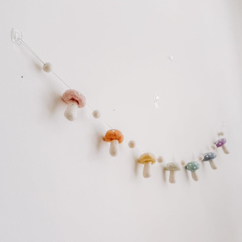 A felt pastel mushroom garland is displayed on a light, neutral wall, evoking a whimsical and cozy atmosphere. The handcrafted felt garland is attached with transparent hooks and features alternating soft pastel wool felt balls and wool felt mushrooms. Each wool felt mushroom, in blush, honey, mint, and lilac hues, has white dots on top.