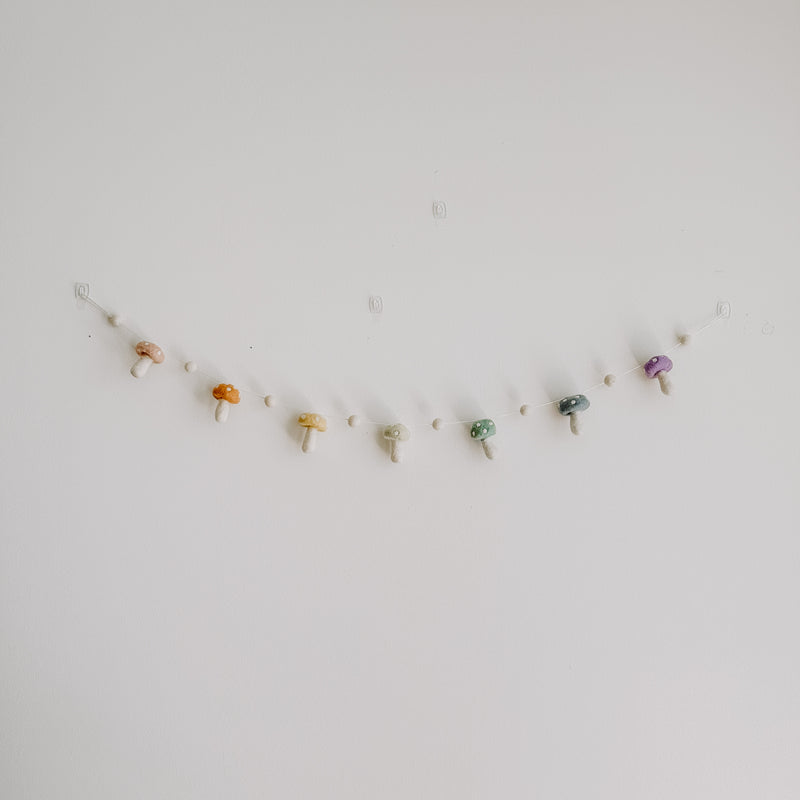 A wool felt pastel mushroom garland is displayed horizontally against a plain white wall, appearing to float. This handmade nursery garland features seven soft wool felt mushrooms in blush, honey, mint, lilac, blue, and orange, each with a spotted cap and creamy white stem. Small, ivory felt balls are evenly spaced between the mushrooms, creating a delicate and charming design.