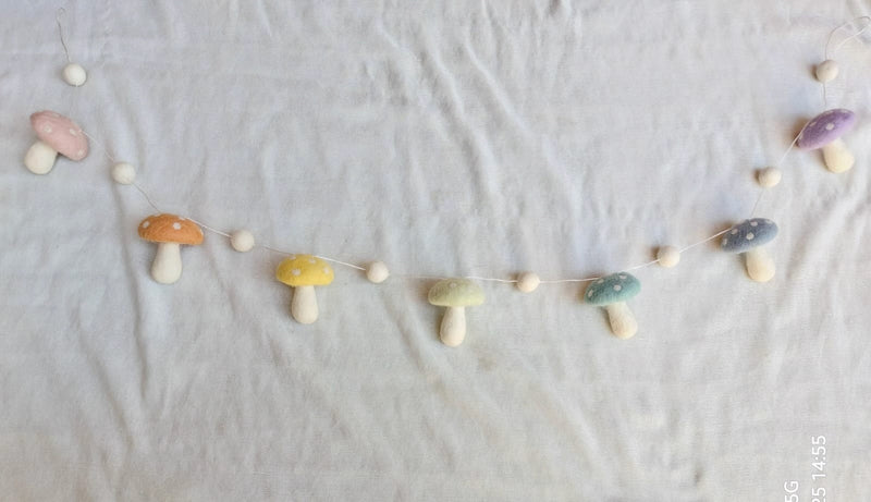 A Wool Felt Pastel Mushroom Garland lies horizontally on a white textured fabric. This charming felt garland showcases handcrafted pastel wool felt mushrooms in pink, orange, yellow, green, and blue, each adorned with white dots on its cap and a white stem. Ivory felt balls are interspersed between the whimsical mushroom shapes.