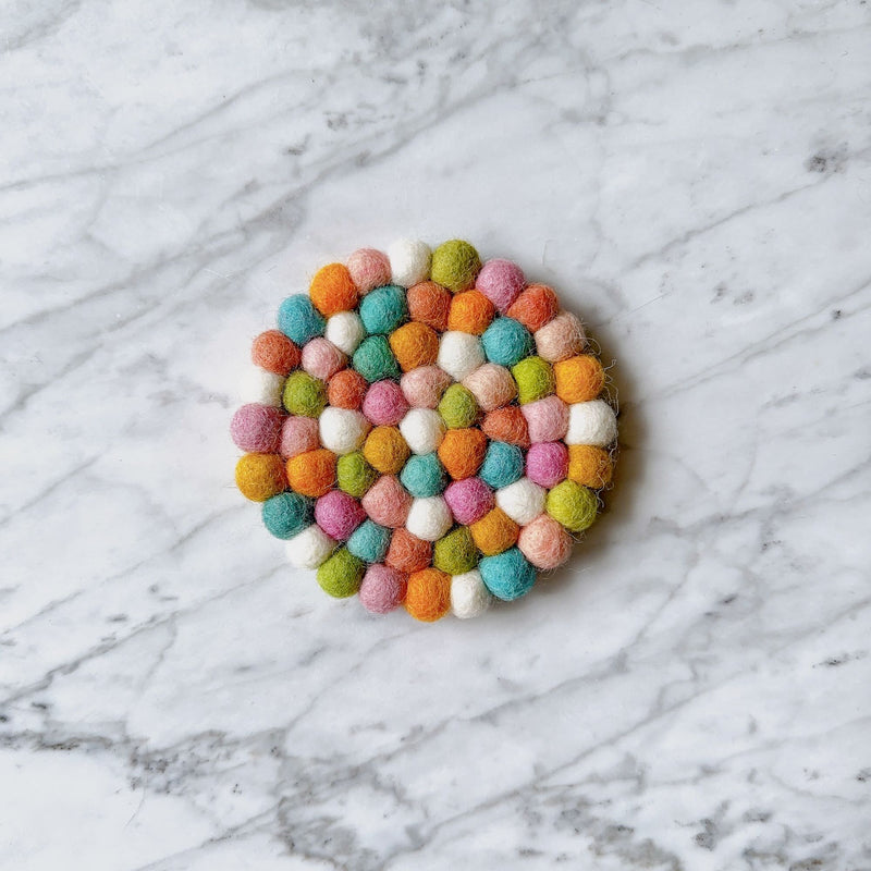 A pastel round felt coaster, part of a set of four, is displayed on a pristine marble backdrop. This handcrafted unique felt coaster is made from individual, approximately half-inch, hand-stitched 100% organic lamb wool felt balls. It showcases a gentle mix of white, green, orange, pink, and blue hues, ethically made by Nepalese female artisans.