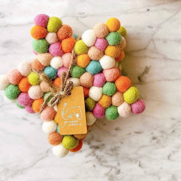 A stack of colorful pastel felt ball star coasters rests on a light-colored marble surface, bundled with a rustic jute string. These circular, handcrafted felt coasters, made from 100% organic lamb wool, feature densely arranged half-inch felt balls in vibrant orange, pink, green, and white. Ethically handmade by Nepalese female artisans, this unique star coaster set offers a tactile and environmentally friendly addition to home decor.