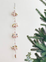 The wool felt pastel unicorn garland is displayed against a clean white wall, positioned beside an artificial Christmas tree. This charming garland features four skillfully needle-felted 3D unicorns, each with a soft white body, pink hooves, and a fluffy multicolored pastel mane. They are connected by a string interspersed with alternating pink and white felt balls, creating a whimsical and festive atmosphere.