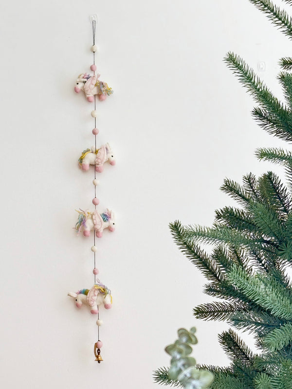 The wool felt pastel unicorn garland is displayed against a clean white wall, positioned beside an artificial Christmas tree. This charming garland features four skillfully needle-felted 3D unicorns, each with a soft white body, pink hooves, and a fluffy multicolored pastel mane. They are connected by a string interspersed with alternating pink and white felt balls, creating a whimsical and festive atmosphere.