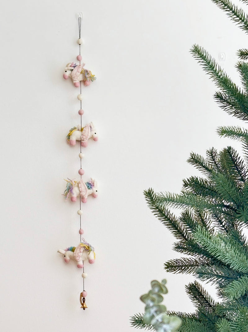 The wool felt pastel unicorn garland is displayed against a clean white wall, positioned beside an artificial Christmas tree. This charming garland features four skillfully needle-felted 3D unicorns, each with a soft white body, pink hooves, and a fluffy multicolored pastel mane. They are connected by a string interspersed with alternating pink and white felt balls, creating a whimsical and festive atmosphere.