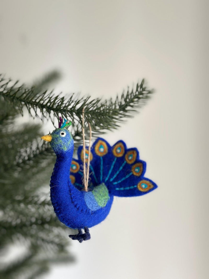A vibrant felt peacock ornament is nestled among green Christmas tree needles, set against a soft white background. This handcrafted felt ornament features a rich blue body, a yellow beak, and a fanned tail adorned with blue and gold accents. Tufts of green, blue, and purple feathers crown its head, while a jute string is attached for hanging.