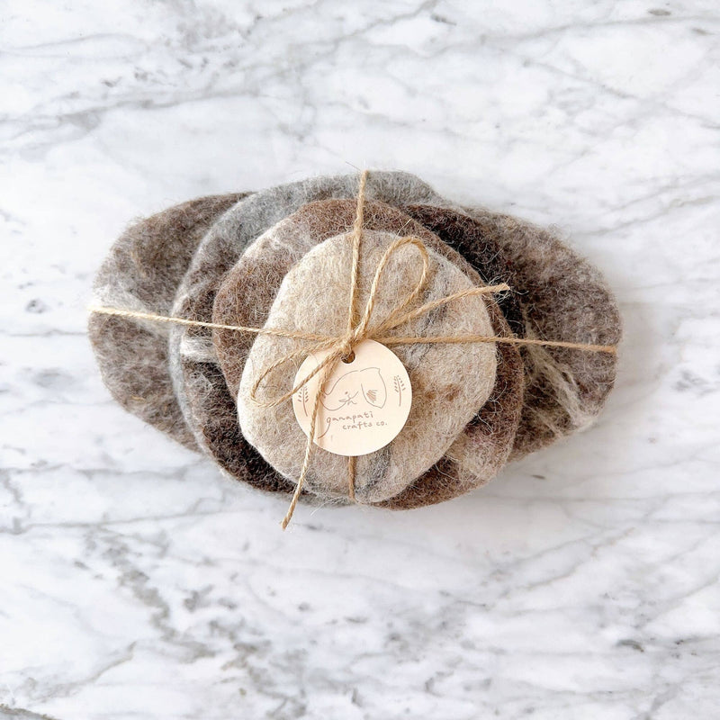This set of five handcrafted wool felt pebble stone coasters is displayed on a light marble surface. Each unique, irregularly shaped coaster is made from organic wool felt in varying earthy tones, evoking rustic charm. The set is beautifully bundled with natural hemp string and features a simple logo tag, highlighting its fair-trade craftsmanship by Nepalese female artisans.