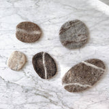 A set of five wool felt pebble stone coasters rest scattered on a white marble surface. Handcrafted from 100% organic lamb wool, these unique felt coasters vary in irregular sizes and shapes, resembling natural pebbles. Their colors range from light beige to dark brown and gray, highlighting their natural texture and eco-friendly design.