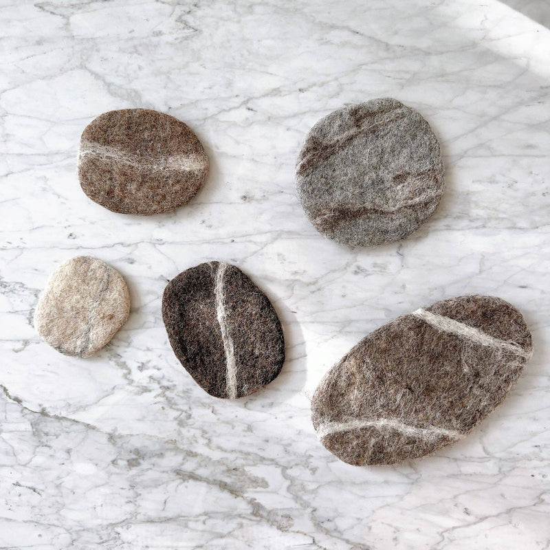 A set of five wool felt pebble stone coasters rest scattered on a white marble surface. Handcrafted from 100% organic lamb wool, these unique felt coasters vary in irregular sizes and shapes, resembling natural pebbles. Their colors range from light beige to dark brown and gray, highlighting their natural texture and eco-friendly design.