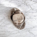 A set of five felt pebble stone coasters rests on a marble surface, stacked to showcase their unique design. These ethically handcrafted wool felt coasters feature irregular sizes and a rough texture, in varying shades of beige and brown. Made from 100% organic lamb wool felt, they offer a rustic and natural aesthetic.