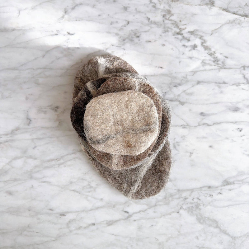 A set of five felt pebble stone coasters rests on a marble surface, stacked to showcase their unique design. These ethically handcrafted wool felt coasters feature irregular sizes and a rough texture, in varying shades of beige and brown. Made from 100% organic lamb wool felt, they offer a rustic and natural aesthetic.