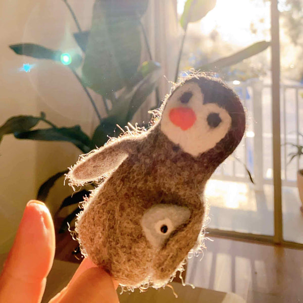 A handmade, needle felted penguin finger puppet, designed to carry a white fish, is gently held by a hand in a warm, softly lit setting. This fair-trade wool felt toy is predominantly brown and cream, featuring a black head, white belly, bright orange beak, and two wings. Its detailed, three-dimensional design stands out against a blurry background of natural window light and greenery.