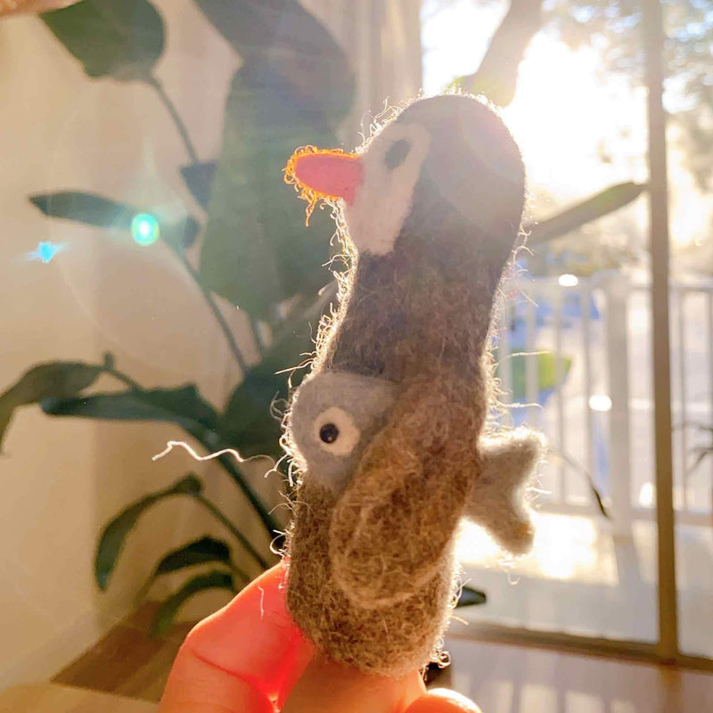 A cheerful, handmade felt penguin carrying a fish finger puppet is held by a female hand. The unique wool felt finger puppet, needle-felted in grey and brown, features a white and pink beak and proudly carries a small fish, presented against a brightly lit, soft-focus background of greenery and sunlight.