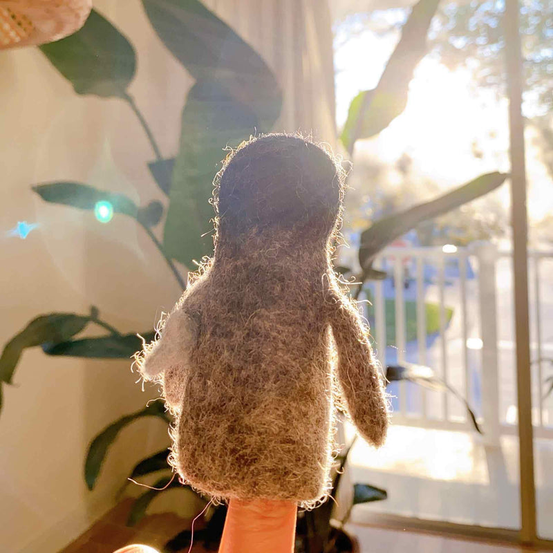 A needle felted penguin finger puppet, carrying a fish, is held on a finger against a bright, sunlit background that includes a plant and window, creating a cozy scene. This handmade wool felt penguin is crafted from textured brown 100% wool felt, featuring small wings and a 3D design visible from all angles.