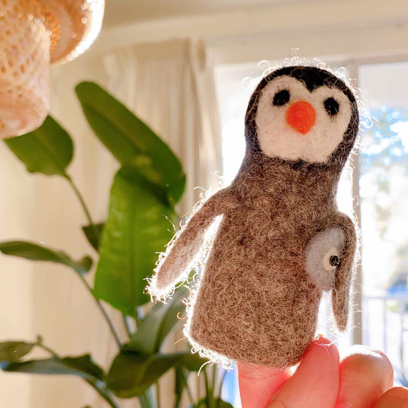 A handcrafted wool felt penguin finger puppet carrying a fish is playfully held by a hand, set against a bright, soft-focus background of a green plant and window. This charming needle felted penguin features a white face with black eyes, an orange beak, and a gray body. Handmade from 100% wool, this unique finger puppet brings imaginative play to life.