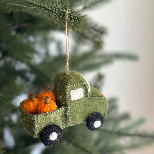 A green felt pickup truck ornament, filled with vibrant orange felt pumpkins in its bed, hangs from a Christmas tree branch. This felt ornament features a white felt window, black wheels with white centers, and is suspended by a jute string against a blurred, light-colored wall.
