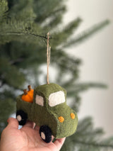 A playful Felt Pickup Truck with Pumpkins Ornament, crafted from green felt, is gently presented by a hand against a festive Christmas tree. The truck features a textured body, white windows, yellow front lights, and black tires with white dots. Orange felt pumpkins fill its bed, and a jute string is attached for hanging, adding rustic charm.