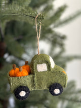 A green felt pickup truck with pumpkins ornament hangs playfully on a Christmas tree. This festive felt ornament features a textured green surface, white felt windows, and black wheels with white centers. Its bed is loaded with several orange felt pumpkins with brown stems, adding an autumnal charm. A jute string is attached for easy hanging.