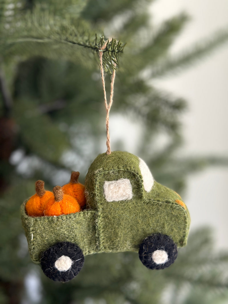 A green felt pickup truck with pumpkins ornament hangs playfully on a Christmas tree. This festive felt ornament features a textured green surface, white felt windows, and black wheels with white centers. Its bed is loaded with several orange felt pumpkins with brown stems, adding an autumnal charm. A jute string is attached for easy hanging.