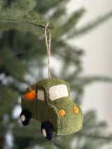 A charming handmade felt pickup truck ornament, packed with bright orange felt pumpkins, hangs against a softly blurred background of green pine needles. The textured green felt truck features white felt windows and orange headlights. Its black wheels have white centers, and a jute string is attached for hanging, creating a cozy, seasonal look.