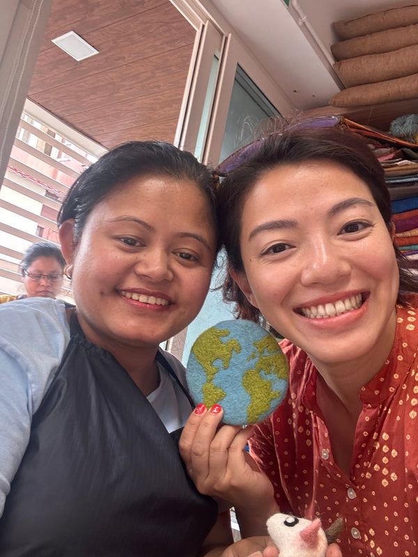 A round felt Planet Earth coaster is held by two smiling women in a bright, cheerful setting. The organic lamb wool felt coaster features green continents against a blue ocean backdrop, with visible stitching highlighting its ethically handcrafted quality. These unique, eco-friendly coasters evoke global awareness.