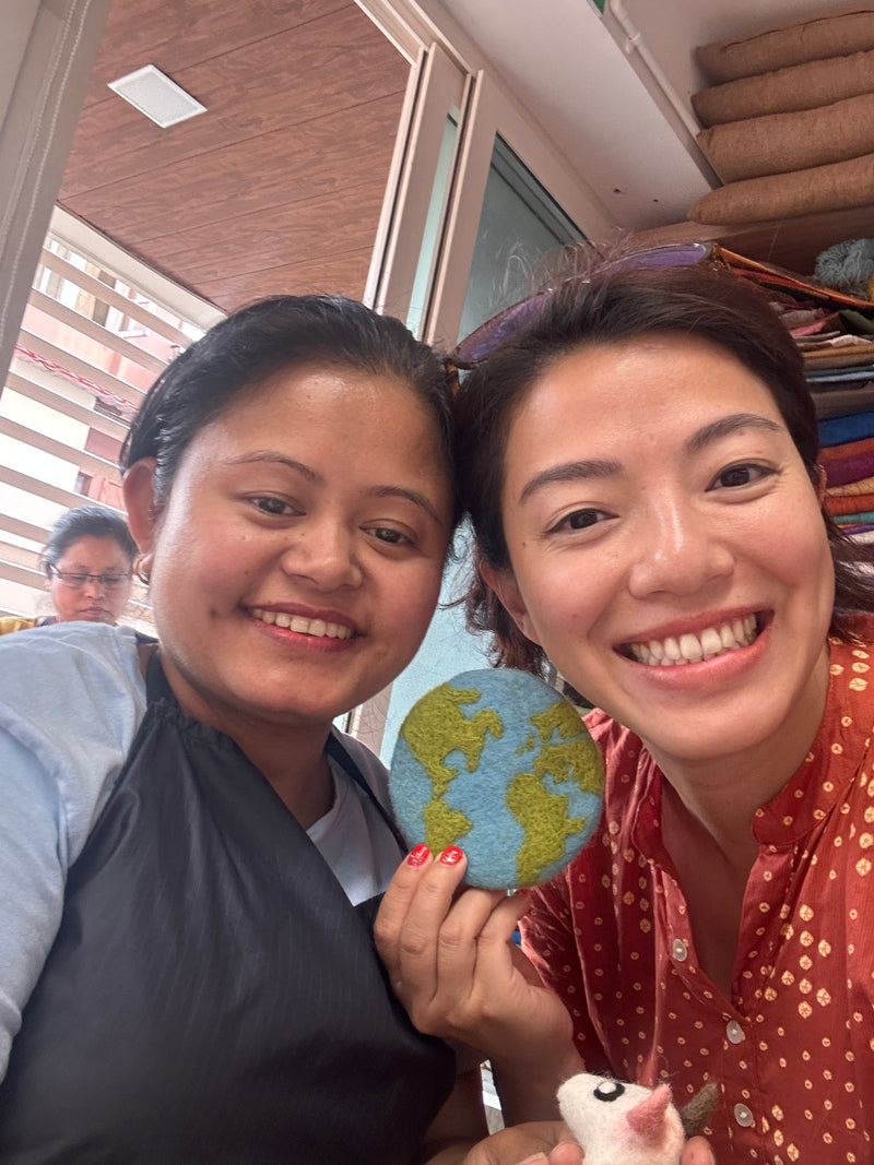A round felt Planet Earth coaster is held by two smiling women in a bright, cheerful setting. The organic lamb wool felt coaster features green continents against a blue ocean backdrop, with visible stitching highlighting its ethically handcrafted quality. These unique, eco-friendly coasters evoke global awareness.