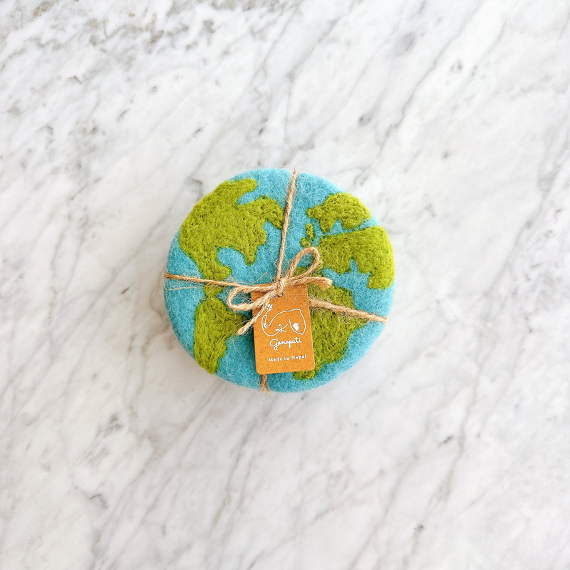 A set of four organic wool felt Planet Earth coasters rests neatly bundled on a white marble surface. Each round, handcrafted felt coaster features a light blue ocean base with green continents, evoking a global design. The ethically made set is tied with a rustic jute string bow and a small 'Ganapati Made in Nepal' tag, highlighting its fair-trade craftsmanship.
