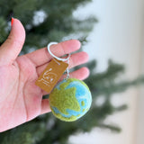 A wool felt planet keychain, ideal for Earth Day, is gently held by a female hand against a blurred green tree background. This round, needle felted keychain showcases green landmasses, blue oceans, and white gradient tie-dye water on its map-inspired design, attached to a metal key ring.