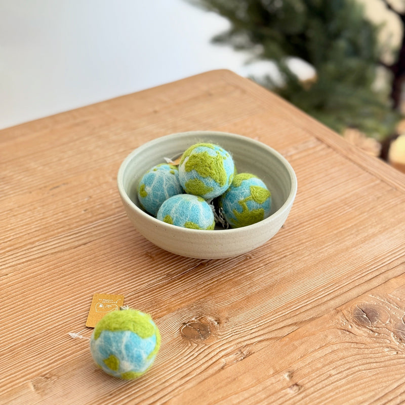 Multiple wool felt planet keychains are displayed on a rustic wooden surface, some nestled within a light grey ceramic bowl, creating an eco-conscious display. These round, handcrafted felt keychains feature a white gradient tie-dye for water and light green for landmasses, all with a textured, needle-felted finish. One planet felt keychain rests outside the bowl, showcasing its attachment.