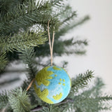 A spherical Felt Planet ornament, crafted from 100% organic lamb wool, hangs from a verdant pine tree branch. This needle felted ornament features a map-like design with white gradient tie-dye for water and green felt continents, creating a textured, handcrafted appearance. A jute string allows it to hang naturally, embodying an eco-friendly and sustainable charm.
