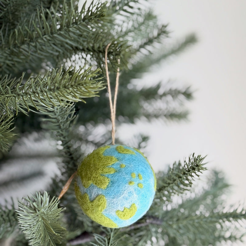 A spherical Felt Planet ornament, crafted from 100% organic lamb wool, hangs from a verdant pine tree branch. This needle felted ornament features a map-like design with white gradient tie-dye for water and green felt continents, creating a textured, handcrafted appearance. A jute string allows it to hang naturally, embodying an eco-friendly and sustainable charm.