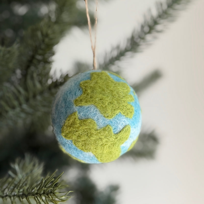 A spherical felt planet ornament hangs gracefully on a green Christmas tree, set against a bright, airy background. This needle-felted wool felt ornament features white gradient tie-dye oceans and green land masses, with a slightly fuzzy texture. A jute string allows this handcrafted felt ornament to be easily hung, ideal for an eco-conscious display.
