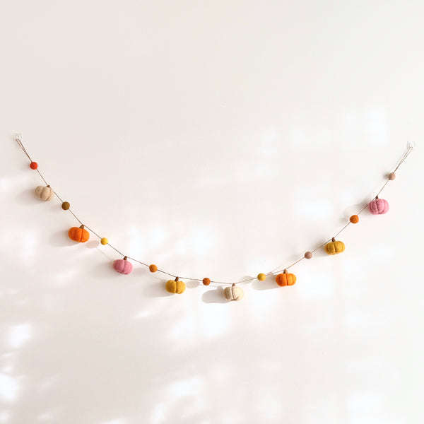 A handcrafted wool felt pumpkin garland is displayed, gently curved, against a clean white wall. This 48-inch autumn-themed garland features eight felted pumpkins in various fall colors, including orange, tan, light pink, and golden yellow. The pumpkins are strung together with jute thread, creating a cozy and charming seasonal decoration.