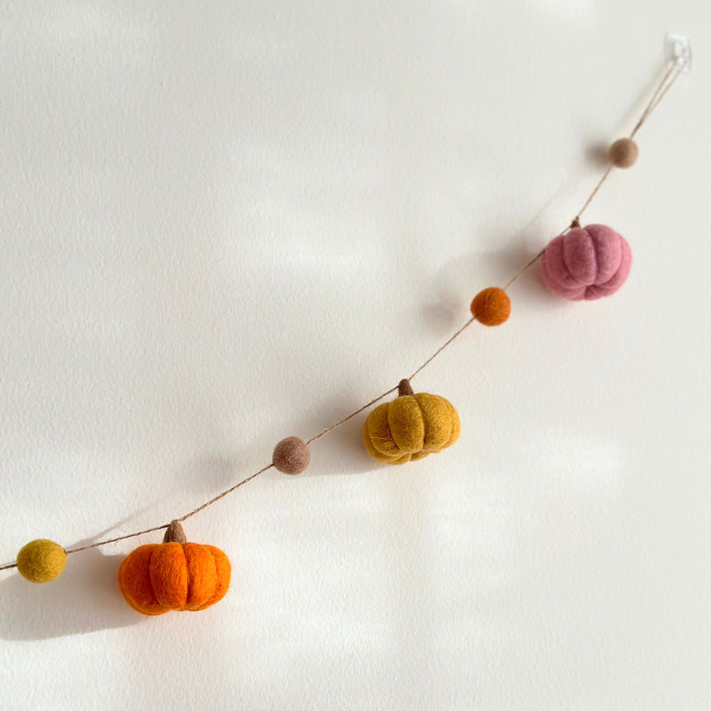 A handcrafted wool felt pumpkin garland hangs against a plain white wall. It features vibrant orange, pink, and yellow felted pumpkins, each with a small brown stem, strung together on a natural jute thread, adding a warm, autumnal touch.