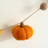 A wool felt pumpkin garland is strung across a plain, off-white backdrop, creating a warm autumn atmosphere. The garland features vibrant orange, yellow, and brown felted wool pumpkins, each with a round, textured body and a detailed brown stem, strung along a visible jute thread.