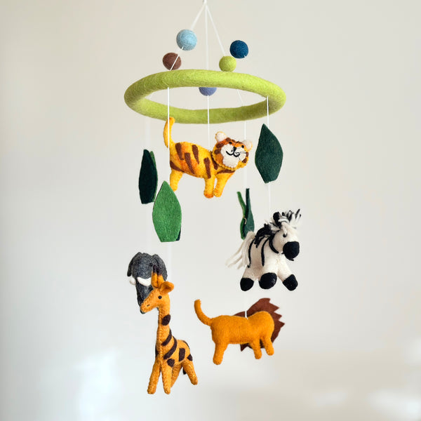 The Felt Safari Animal Baby Mobile is suspended against a plain, off-white background. This whimsical wool felt mobile features a light green ring from which hand-felted animals like a striped tiger, a black and white zebra, a gray elephant with white tusks, a spotted giraffe, and a yellow lion with a brown mane hang. Green leaves and blue, light green, and brown felt balls are interspersed, completing the playful design.