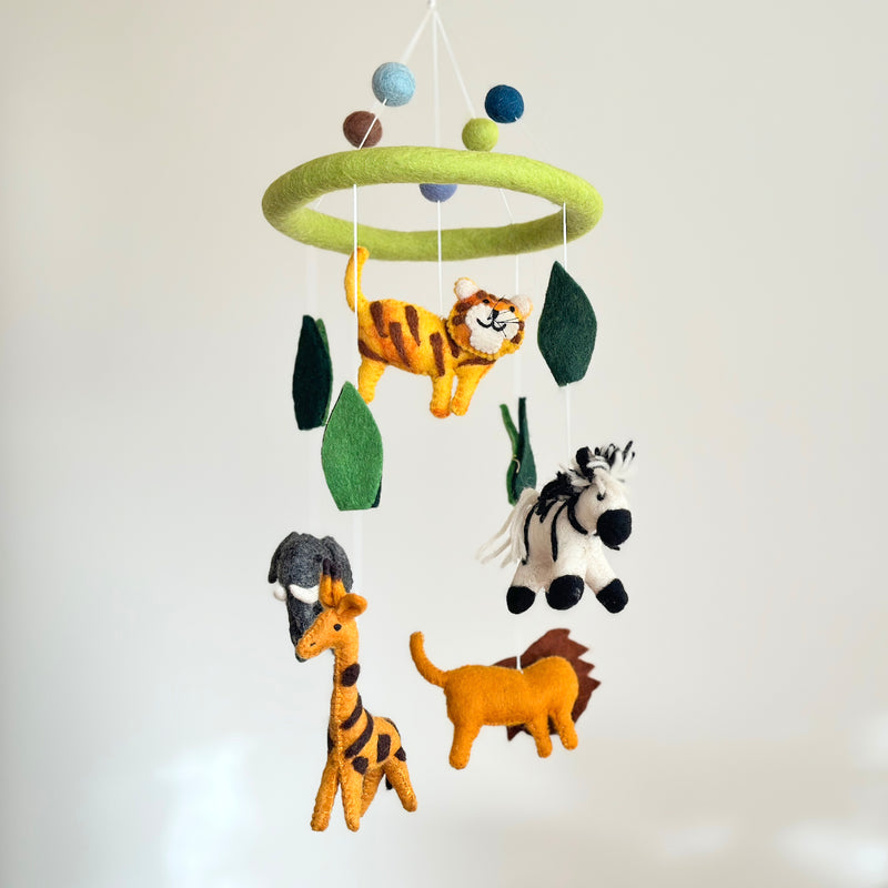 The Felt Safari Animal Baby Mobile is suspended against a plain, off-white background. This whimsical wool felt mobile features a light green ring from which hand-felted animals like a striped tiger, a black and white zebra, a gray elephant with white tusks, a spotted giraffe, and a yellow lion with a brown mane hang. Green leaves and blue, light green, and brown felt balls are interspersed, completing the playful design.