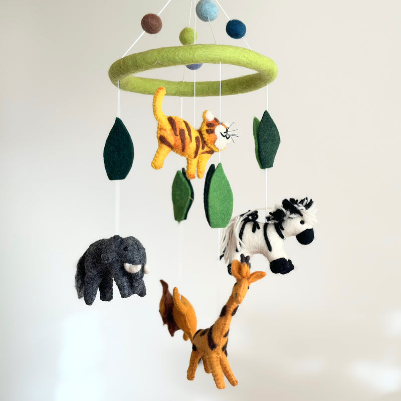 A felt safari animal baby mobile hangs against a plain white background. It features a light green felt ring from which a grey elephant, a black-and-white zebra, an orange-and-black striped giraffe, and an orange-and-black striped tiger are suspended by white strings. Dark green felt leaves and blue, light green, and brown spheres are interspersed above and around the animals. Crafted from soft, hand-felted materials, this baby mobile evokes a calm and playful atmosphere for a nursery.