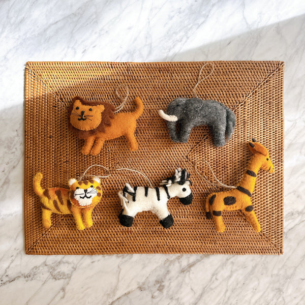 A set of five unique handmade felt safari animal ornaments are playfully arranged on a woven Bali rattan placemat against a white marble backdrop. The set includes a brown-maned lion felt ornament with stitched features, a gray elephant felt ornament with white tusks, a yellow and brown striped tiger felt ornament with a smiling face, a black and white striped zebra felt ornament, and a yellow giraffe felt ornament with brown spots. Each charming wool felt ornament has a jute string for hanging.