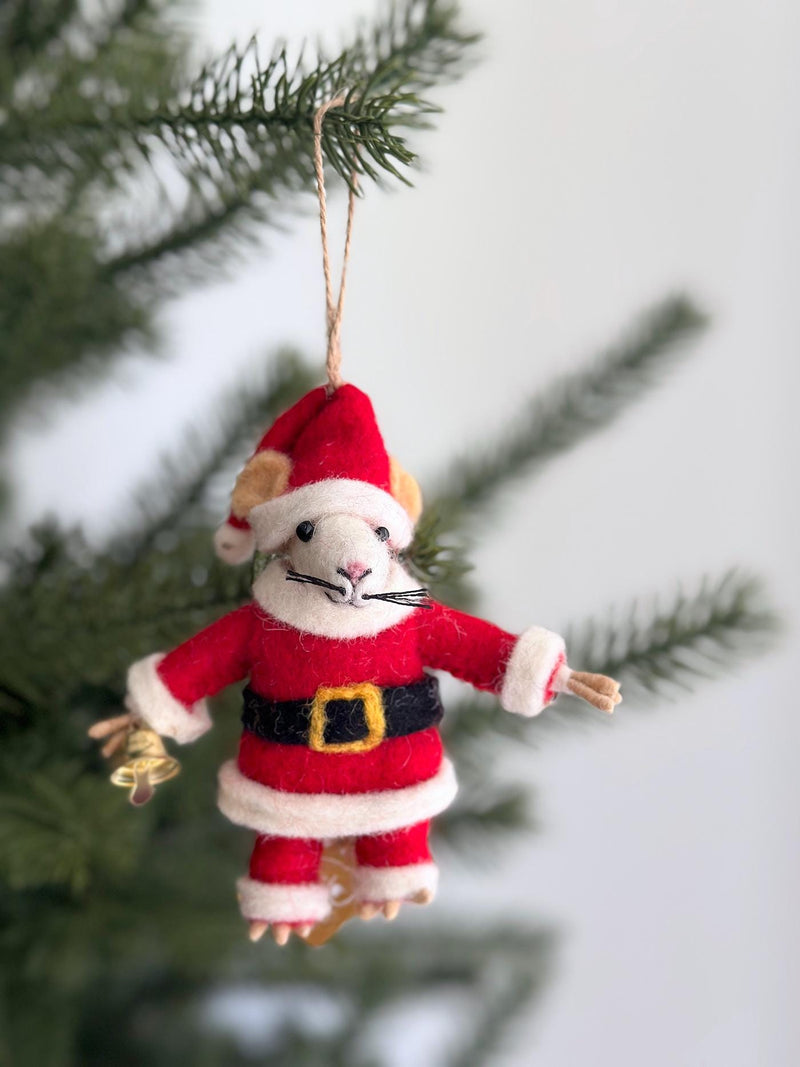 A handmade wool felt Santa mouse ornament hangs from a green Christmas tree branch, evoking a festive and joyful mood. The charming felt mouse wears a red Santa suit with white trim, a black belt with a gold buckle, and a red Santa hat. It features a white face, pink nose, black whiskers, and holds a small gold bell.
